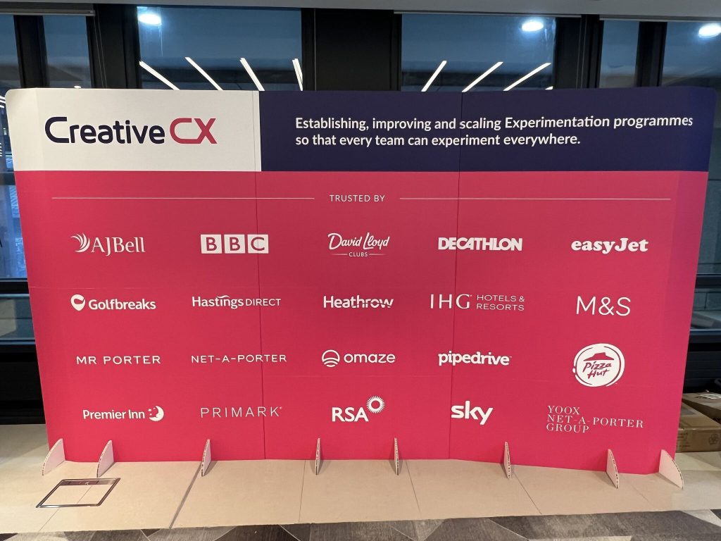 Creative CX conference stand featuring a branded banner with the tagline: Establishing, improving and scaling Experimentation programmes so that every team can experiment everywhere." The banner highlights logos of various brands we work with, including AJ Bell, BBC, Sky, easyJet, and M&S.